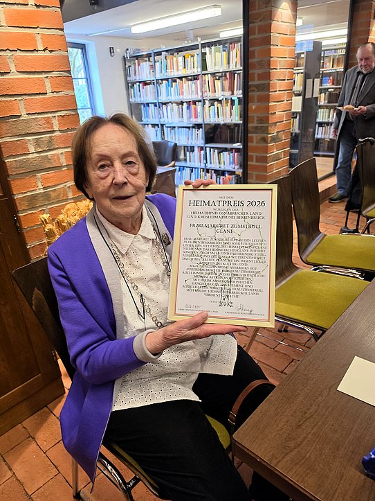 Die Preisträgerin Margret Zumstrull mit der Urkunde - im Bildhintergrund das von ihr aufgebaute Archiv